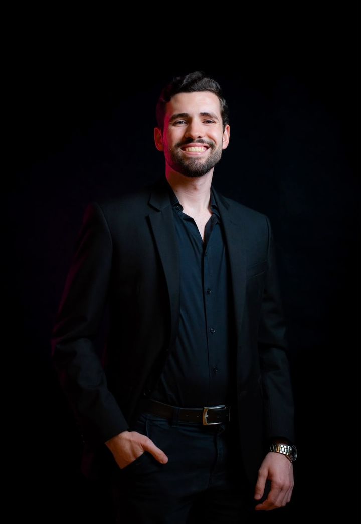 Fernando do Val sorrindo, de terno e camisa pretos, com a mão no bolso, em fundo escuro.